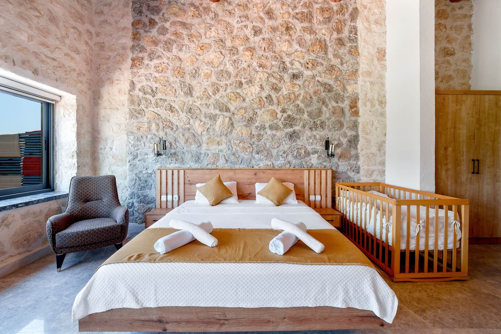 A hotel bedroom with a made bed, stone walls and daylight from a window